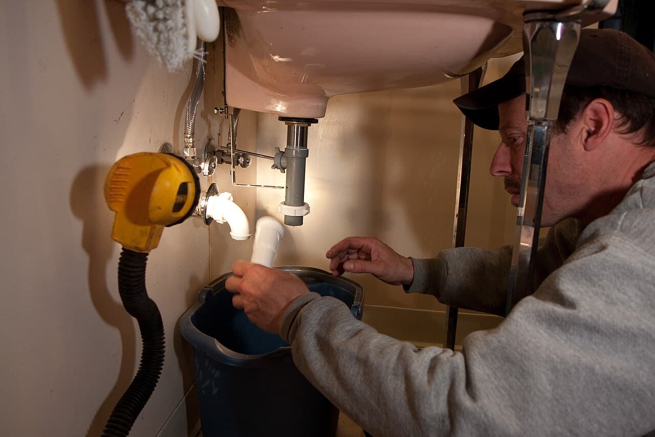 Reference photograph of a plumber working on a disconnected drain beneath a bathroom sink in the United States